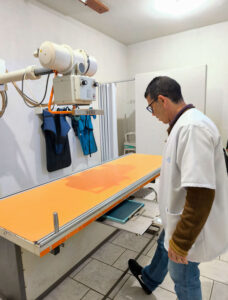 Medical professional in a white coat stands beside an orange radiography table with an overhead X-ray imager in a clinic setting.