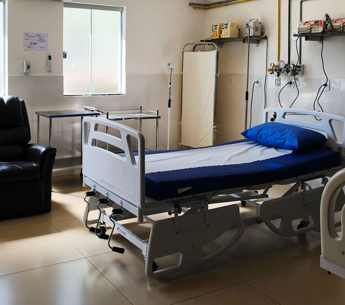 Hospital bed with blue sheets and pillow in a clean clinical room, medical equipment mounted on the wall nearby and a window to the left.